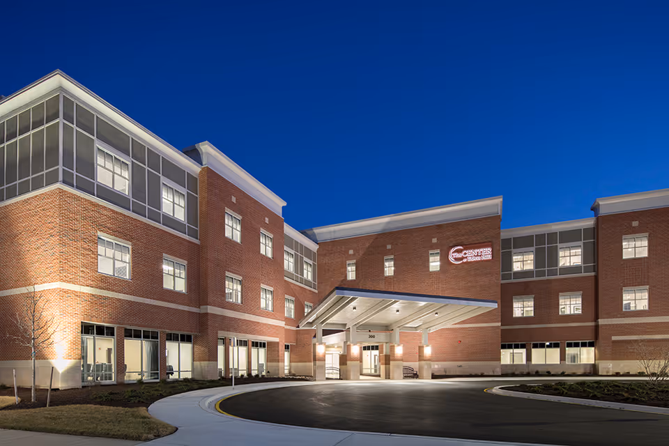 Exterior view of a large, modern brick building at dusk with multiple windows lit from inside. The building has a covered entrance with lights and a circular driveway. A sign on the building reads 'Center At Eden Hill'.