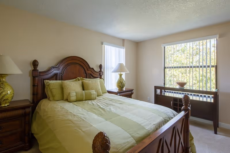 Sunlit bedroom with a wooden bed dressed in green bedding, matching nightstands and lamps, and a window with vertical blinds.