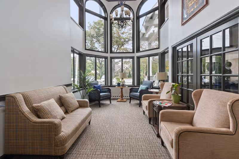 A bright and cozy sitting area with large arched windows letting in natural light. The room features a plaid sofa with matching pillows on the left, two beige armchairs on the right, and two dark blue armchairs near the windows. A small wooden table with a lamp and a plant is placed between the blue chairs, and another side table with a plant and magazines is next to the beige armchairs. The carpet is patterned, and a chandelier hangs from the ceiling.