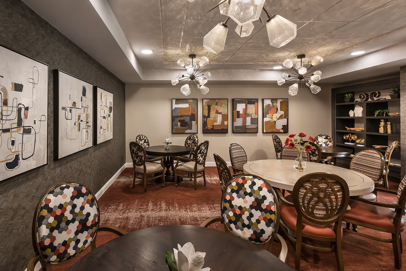 A well-decorated dining room with multiple round and oval tables surrounded by chairs featuring colorful patterned upholstery. The room has modern light fixtures on the ceiling, abstract artwork on the walls, and a shelving unit with decorative items. The carpet is a reddish-brown color, and the overall ambiance is warm and inviting.