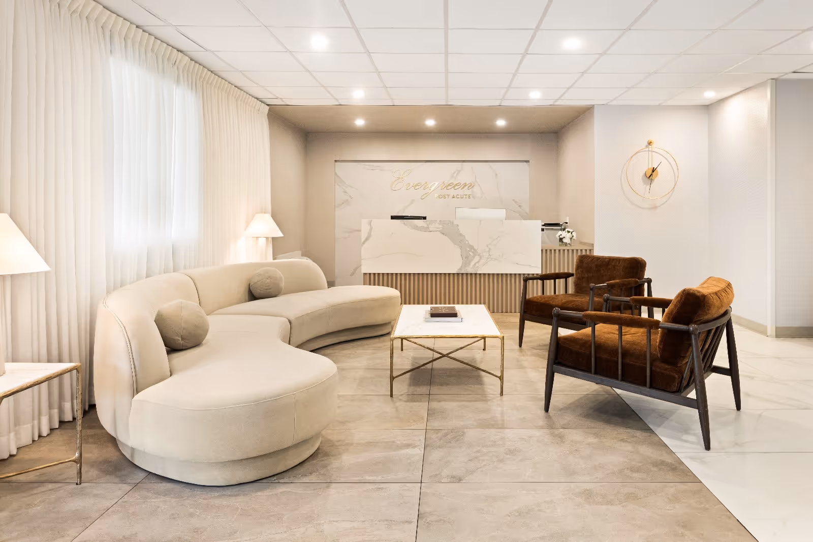 A modern and elegant reception area with a curved beige sofa, two brown cushioned armchairs, a marble coffee table, and a reception desk with the sign 'Evergreen Post Acute' on the wall behind it. The room features large floor tiles, sheer white curtains, and soft lighting.