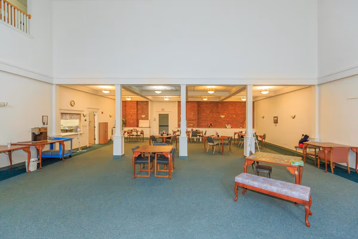 Large communal dining room with scattered tables and chairs, white pillars, and a brick-wall serving area in the back.