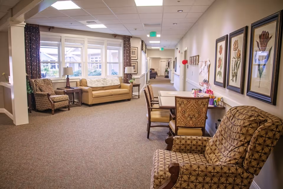 A well-lit hallway in a senior living facility with patterned armchairs, a beige sofa, side tables with lamps, and a table with chairs along the wall. The walls are decorated with framed botanical prints and there are windows letting in natural light.