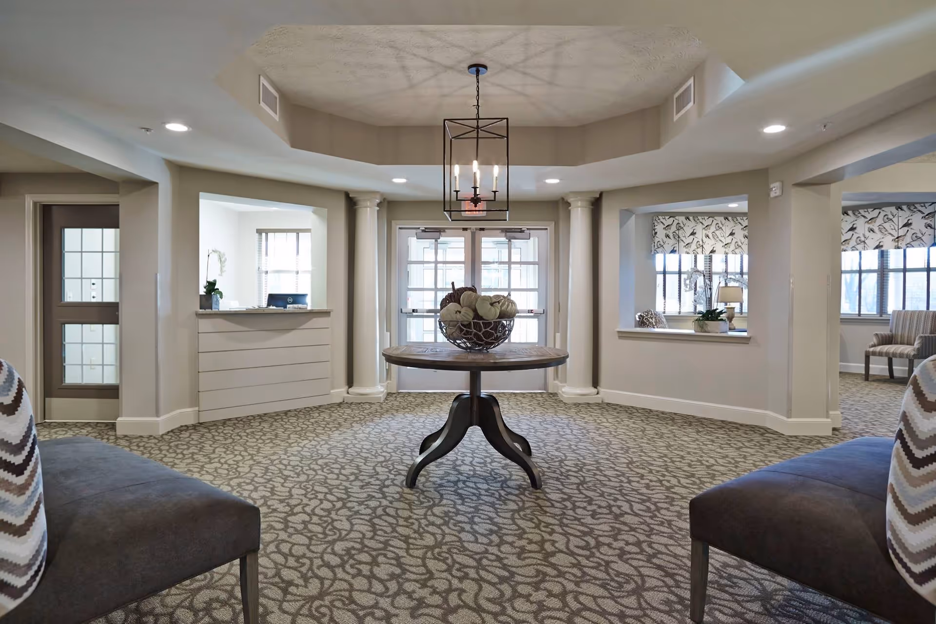 A spacious, well-lit interior common area with patterned carpet, a round wooden table with decorative pumpkins in the center, two upholstered chairs with chevron-patterned cushions on either side, and large windows with floral valances. The area features columns, a chandelier, and a reception desk to the left.