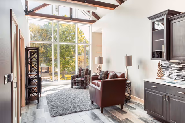 Bright living room with floor-to-ceiling windows, a rug, leather and patterned chairs, side tables with lamps, and kitchenette cabinets.