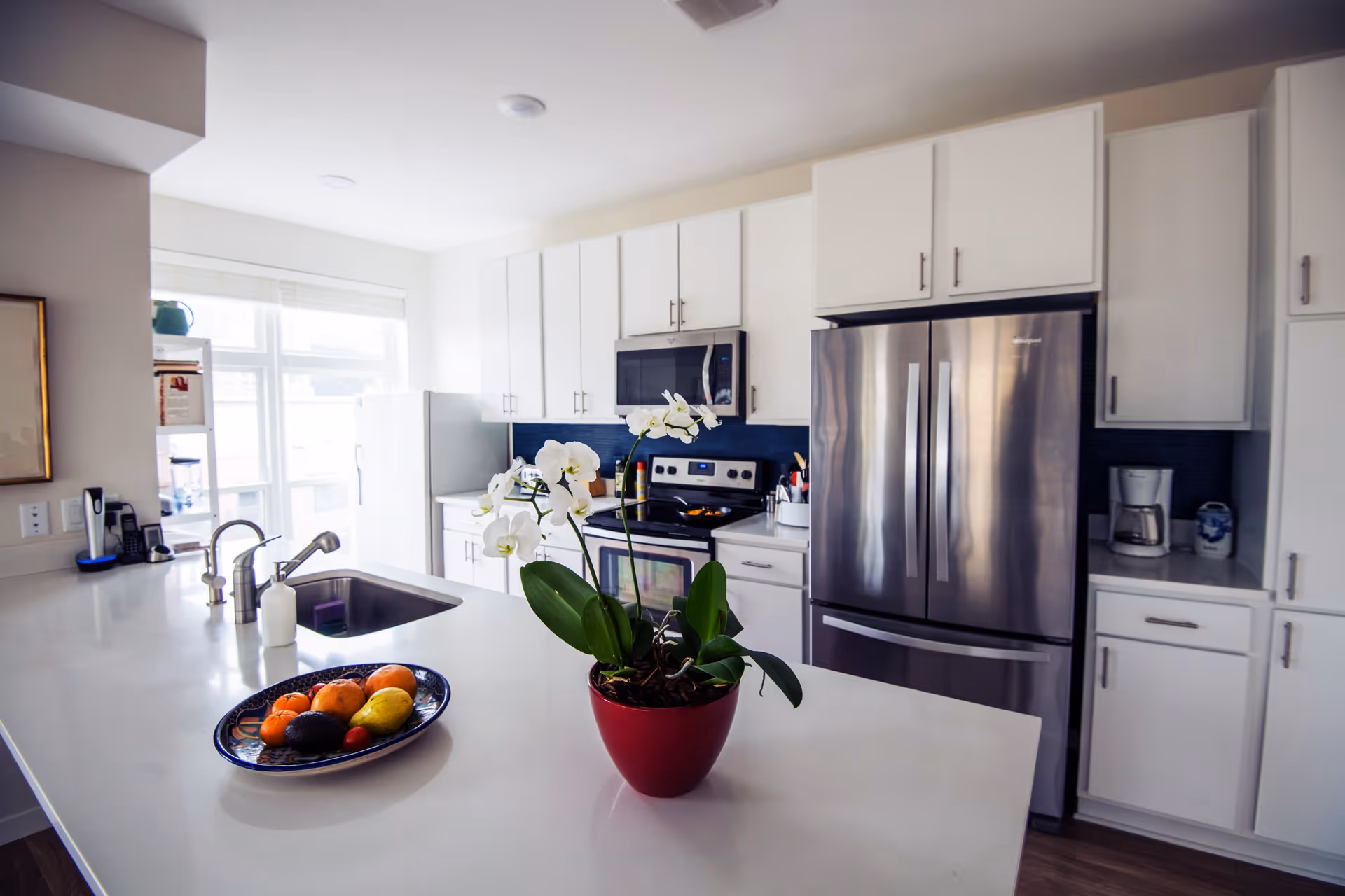 Modern kitchen with white cabinets, stainless steel refrigerator, oven, and microwave. A white countertop island features a red pot with white orchids and a plate of assorted fruits. Large windows provide natural light in the background.