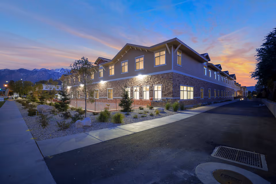 Exterior view of a two-story senior living facility building at dusk with lit windows, landscaped garden area with small trees and rocks, a sidewalk, and mountains in the background under a colorful sky.