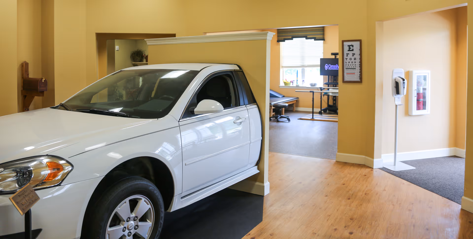 Interior view of a rehabilitation facility room with a white car positioned indoors next to a half wall. The room has yellow walls and wooden flooring. In the background, there is an eye chart on the wall, a hand sanitizer dispenser, a fire extinguisher cabinet, and a desk with a computer monitor and an examination table.
