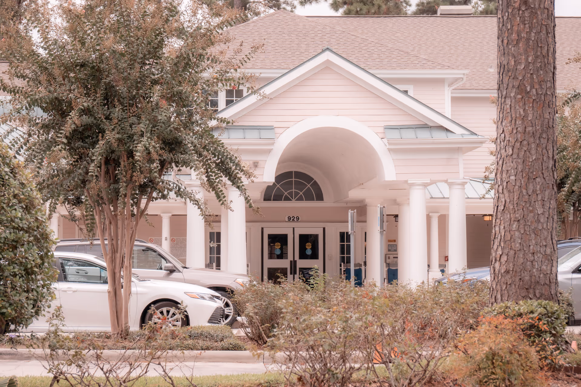 Front entrance of a senior living facility with white columns and an arched roof over the doorway. Several cars are parked in front, and there are trees and shrubs in the foreground.