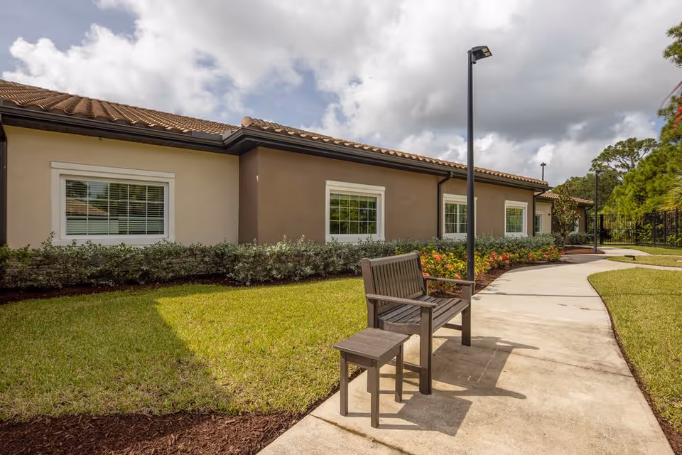Outdoor view of a single-story building with beige and brown walls, white-framed windows, and a tiled roof. A concrete pathway curves alongside the building, bordered by green grass, shrubs, and flowers. A wooden bench and small table are placed on the pathway near a tall lamp post under a partly cloudy sky.