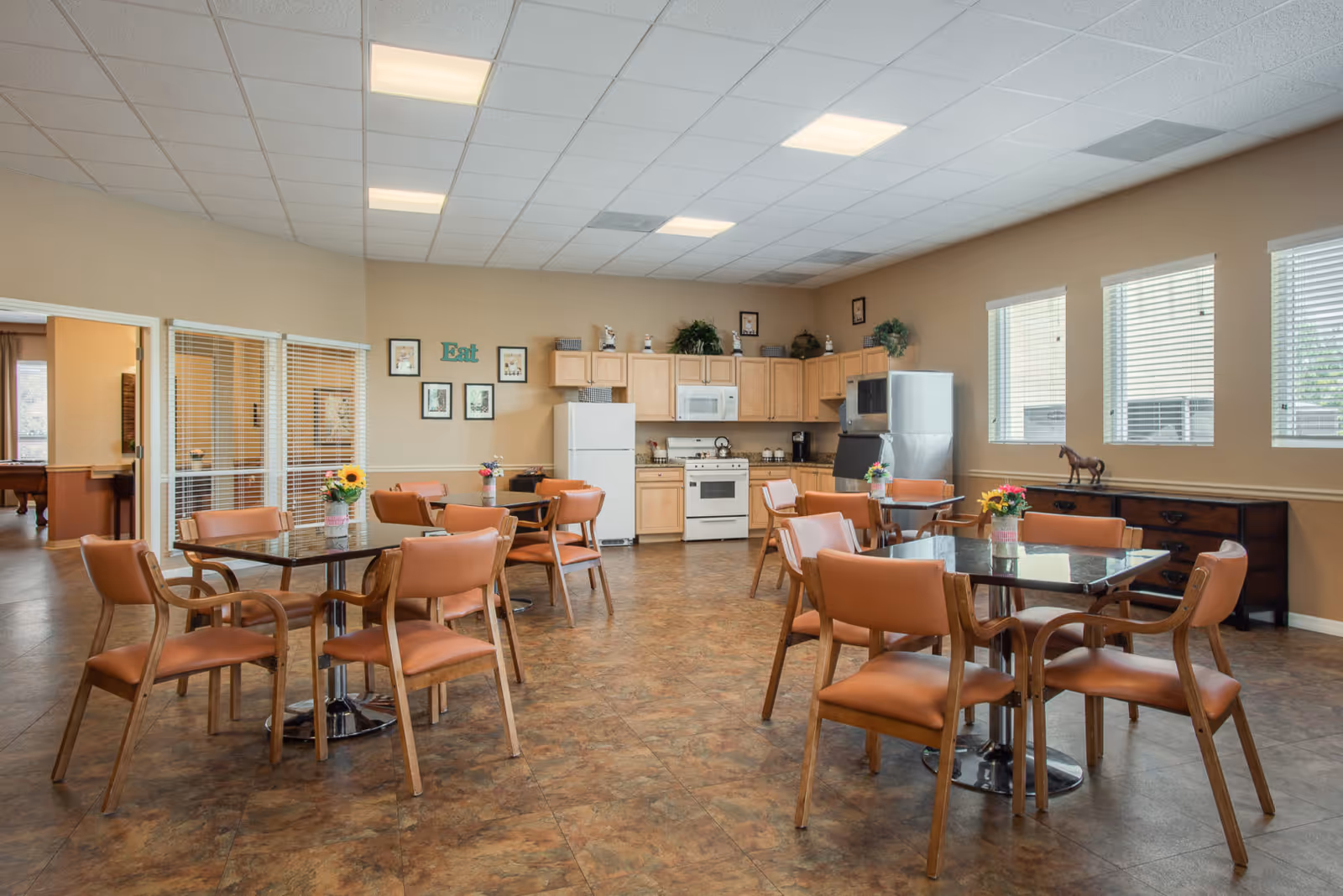 A spacious dining area in a senior living facility with several tables and chairs arranged neatly. The room features a kitchen area with light wood cabinets, a white refrigerator, stove, microwave, and a stainless steel refrigerator. The walls are decorated with framed pictures and the word 'Eat'. Large windows with blinds allow natural light to fill the room. Small flower arrangements are placed on each table.