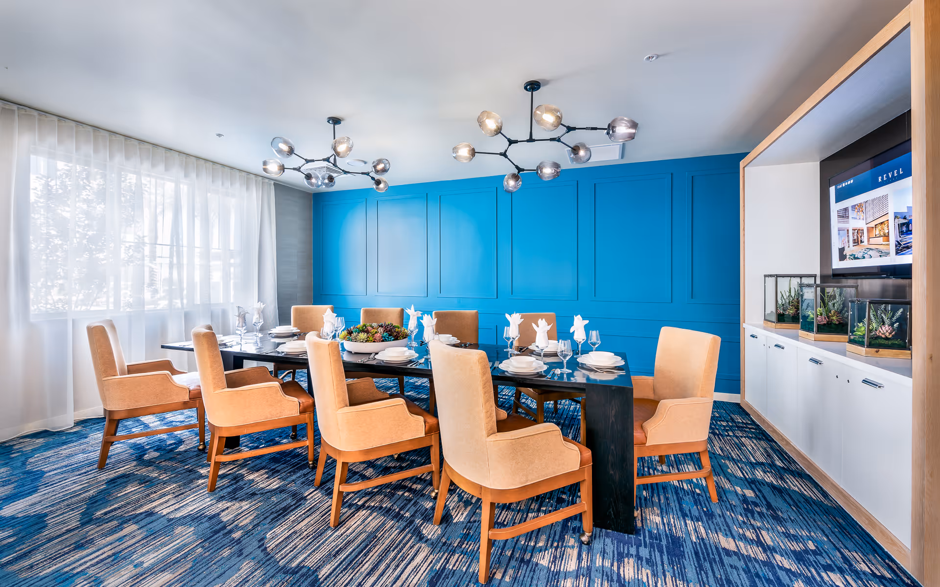 A modern dining room with a long black table set for eight people with beige upholstered chairs. The room features a bright blue accent wall, two contemporary chandeliers, a large window with sheer white curtains, and a built-in cabinet with a TV and decorative plants.
