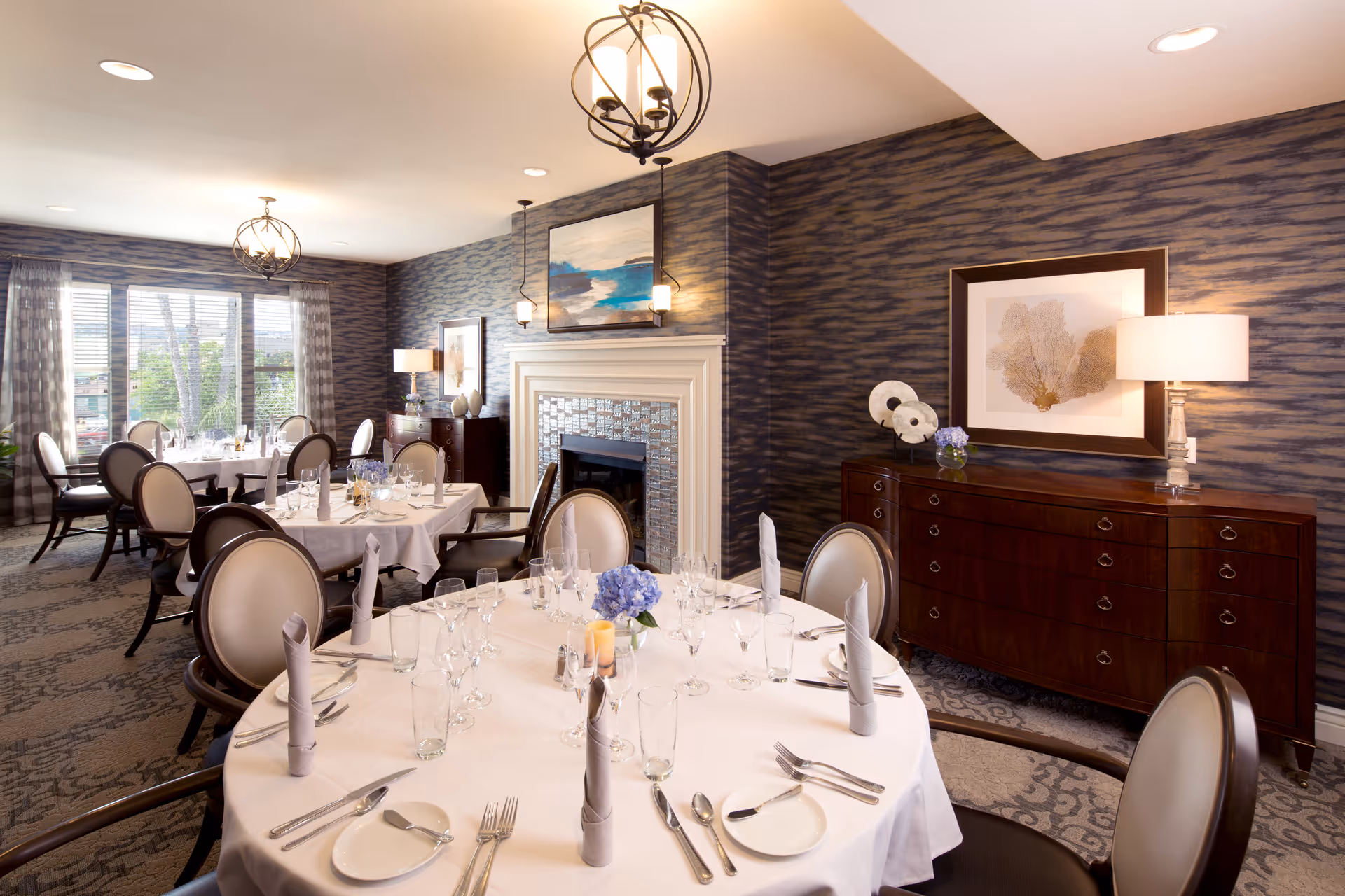 Elegant dining room with round tables covered in white tablecloths, set with glassware, silverware, and napkins. The room features patterned wallpaper, a fireplace with a decorative mantel, framed artwork, and a sideboard with a lamp and decorative items. Large windows with sheer curtains allow natural light to fill the space.