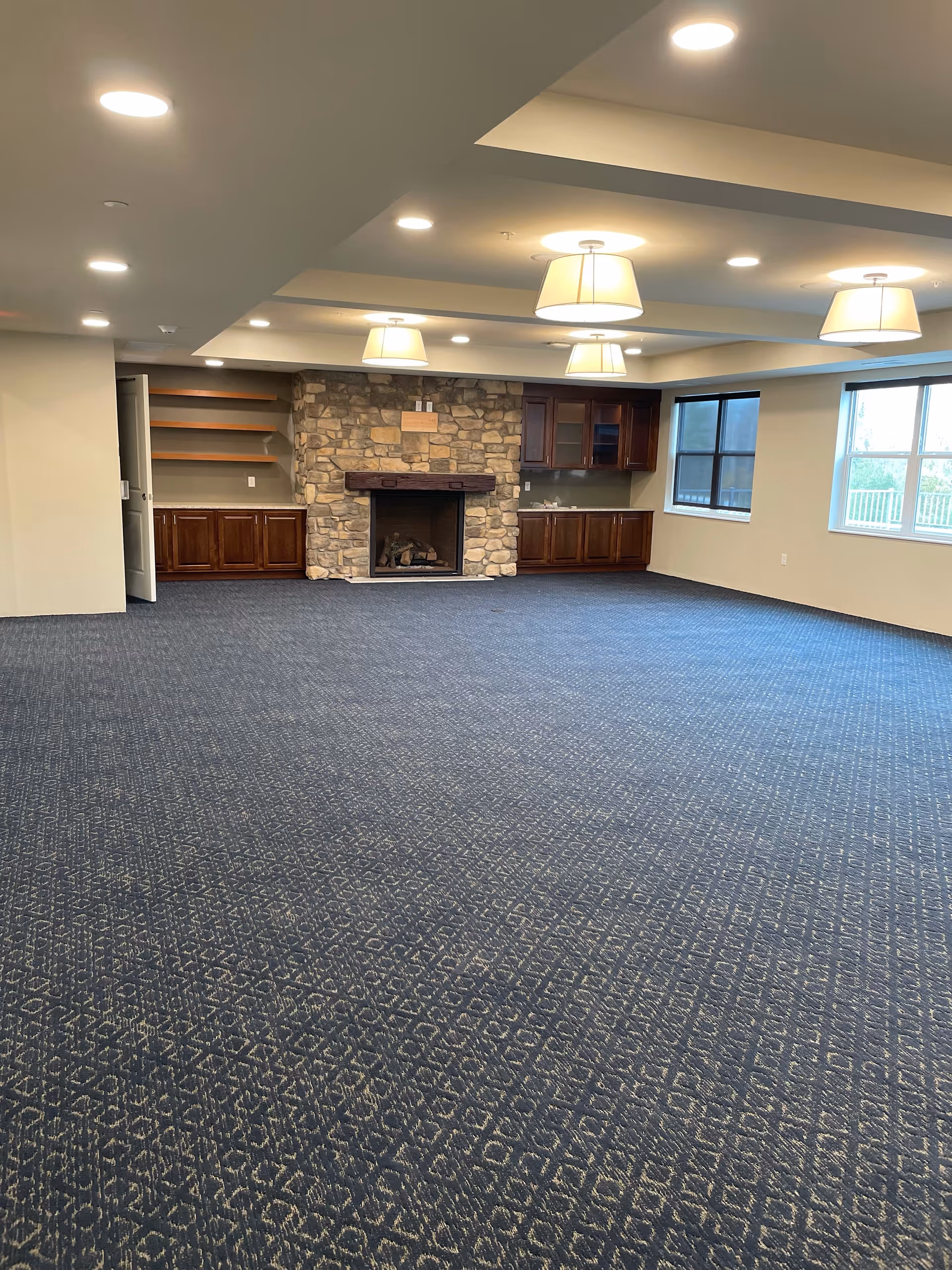 Large carpeted community room with a stone fireplace, built-in cabinets, ceiling lights, and windows.