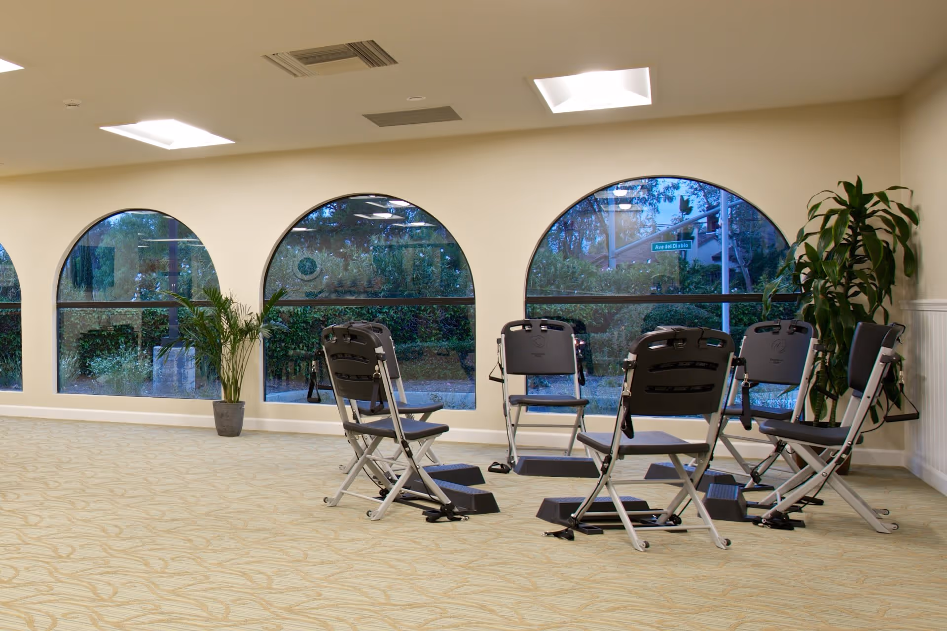 A spacious room with large arched windows showing an outdoor view with greenery. Inside, there are six black folding chairs arranged in a circle with small black footrests in front of each chair. Two potted plants are placed near the windows, and the room has beige walls and carpeted flooring with a subtle pattern.