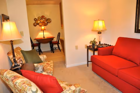 Cozy living room with patterned armchairs, a red sofa, table lamps, and a small dining area visible through a doorway.