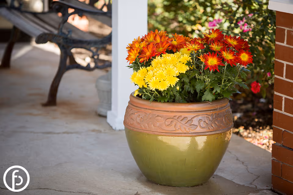 A large decorative ceramic pot with orange, yellow, and red flowers placed on a concrete porch near a white column and a brick wall. A metal bench is partially visible in the background.