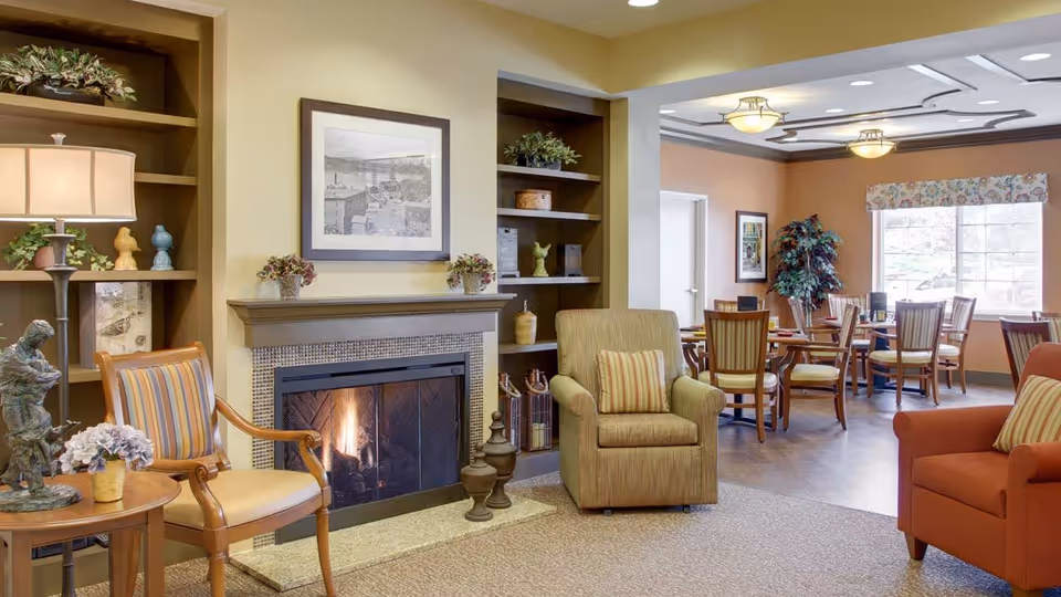 A cozy senior living facility common area featuring a lit fireplace with a framed picture above it, flanked by built-in shelves with decorative items. There are comfortable chairs with striped cushions, a small round table with a lamp and a sculpture, and a dining area with wooden tables and chairs near a large window with floral valance.
