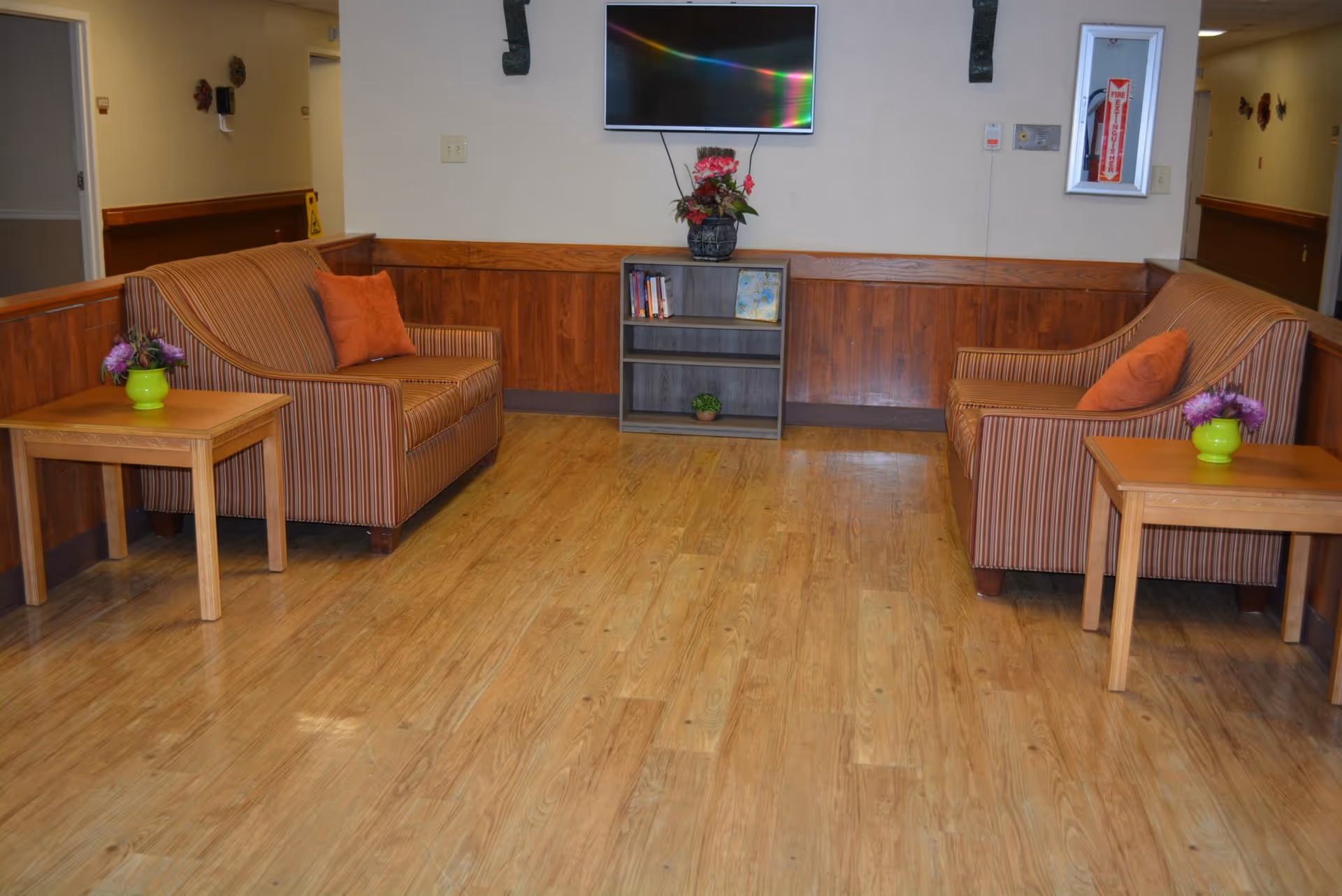 Small seating area with two striped armchairs facing a wall-mounted TV, side tables with flower vases, a small bookshelf, and wood flooring.