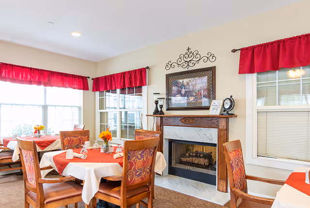 A cozy dining room with several wooden tables covered with white and red tablecloths. Each table has a small floral centerpiece and neatly arranged napkins. The room features a decorative fireplace with a framed picture and ornamental wall decor above it. Large windows with red valances allow natural light to brighten the space.