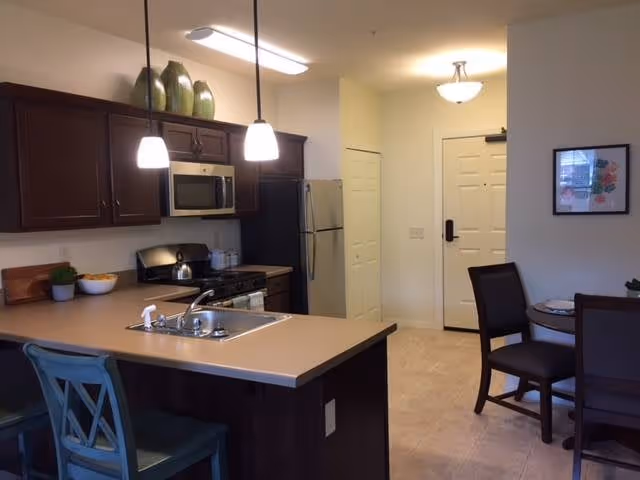 Interior view of a kitchen and dining area in a senior living facility. The kitchen features dark wood cabinets, a stainless steel microwave, stove, and refrigerator. There is a countertop with a sink and two pendant lights hanging above it. Two chairs are placed at the counter. In the background, there is a small dining table with four chairs, a framed picture on the wall, and a white door leading to another room.