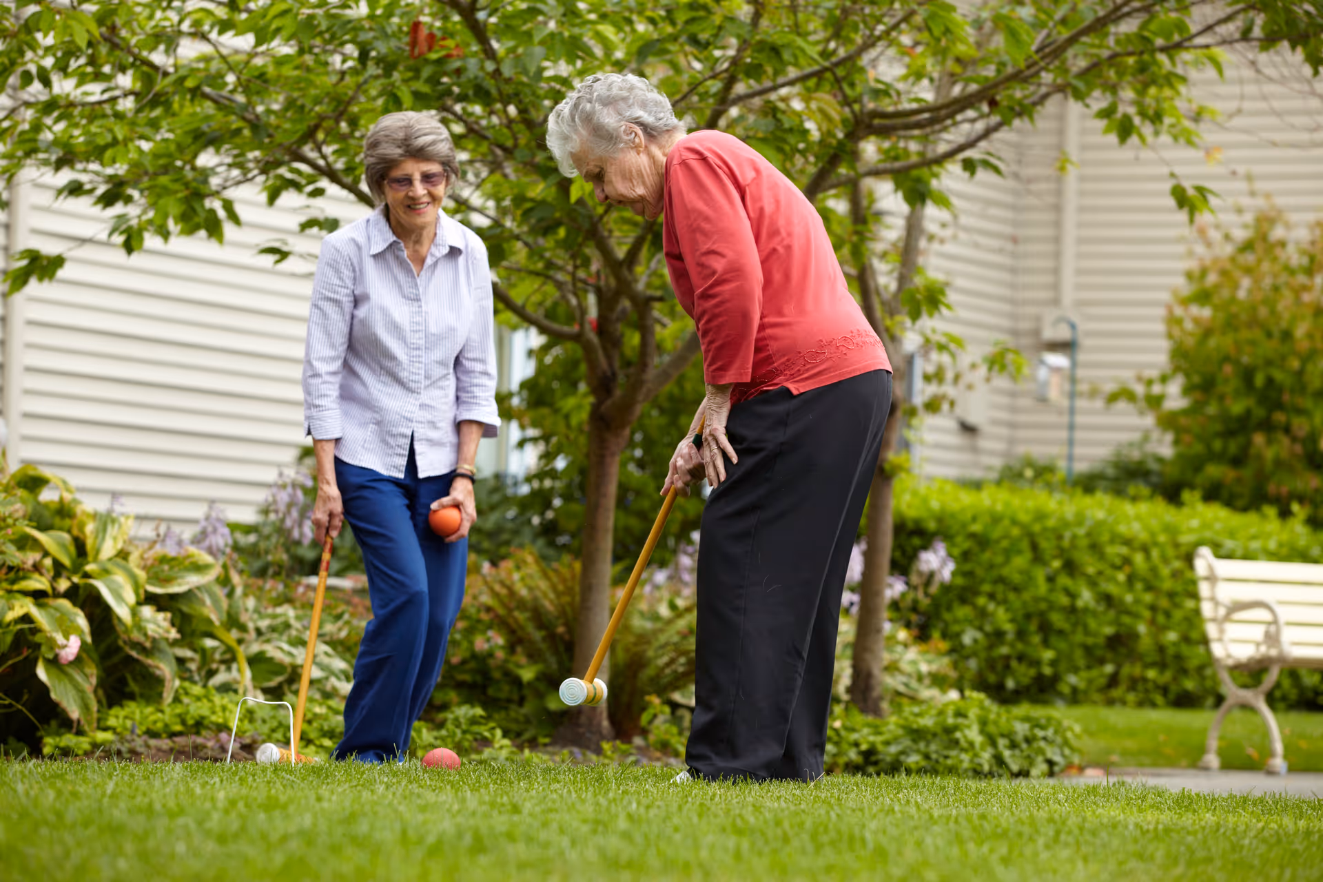 Two elderly women playing croquet on a grassy lawn in a garden area with trees and shrubs, near a building with beige siding and a white bench in the background.