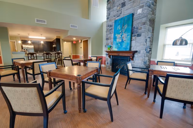 Bright communal dining area with wooden tables and chairs, a stone fireplace with artwork above, and a kitchenette in the background.