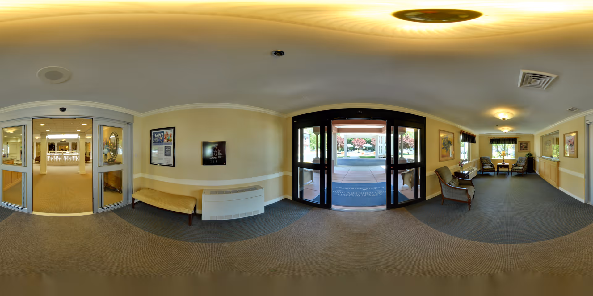 Panoramic view of the entrance lobby of a senior living community. The image shows automatic glass doors leading outside, a bench against the wall, framed artwork, and a seating area with chairs and a table near windows. The space is well-lit with ceiling lights and has a carpeted floor.