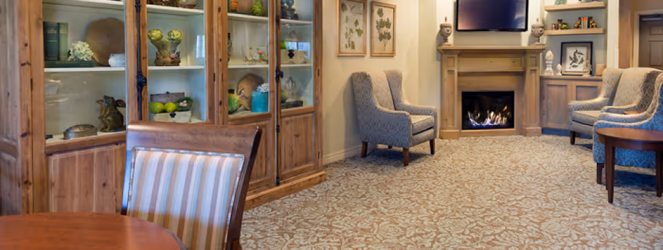 A cozy living room area with a patterned carpet, wooden display cabinets filled with decorative items, several upholstered armchairs, a wooden fireplace with a fire burning, and a flat-screen TV mounted above the fireplace.