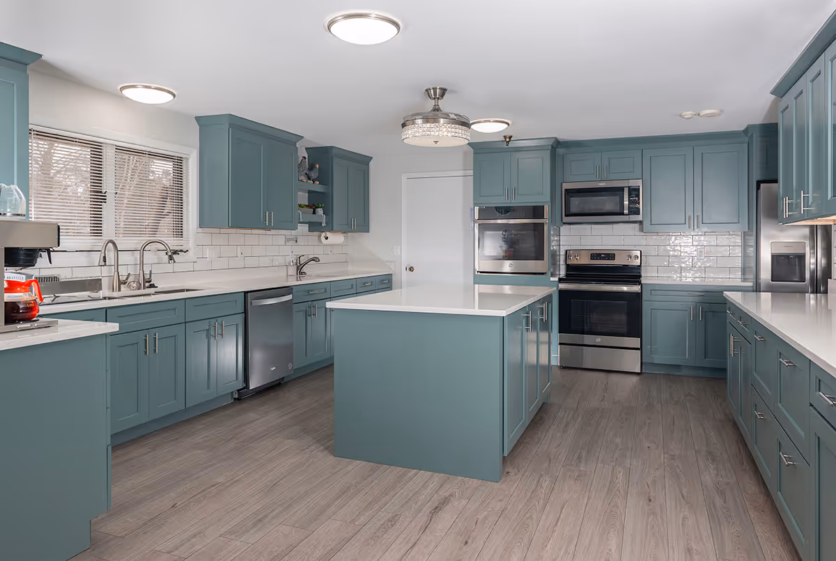 A spacious, modern kitchen with teal cabinets, white countertops, and stainless steel appliances including a dishwasher, oven, microwave, and refrigerator. There is a kitchen island in the center and wood flooring throughout. The room is well-lit with ceiling lights and a window with blinds above the sink.