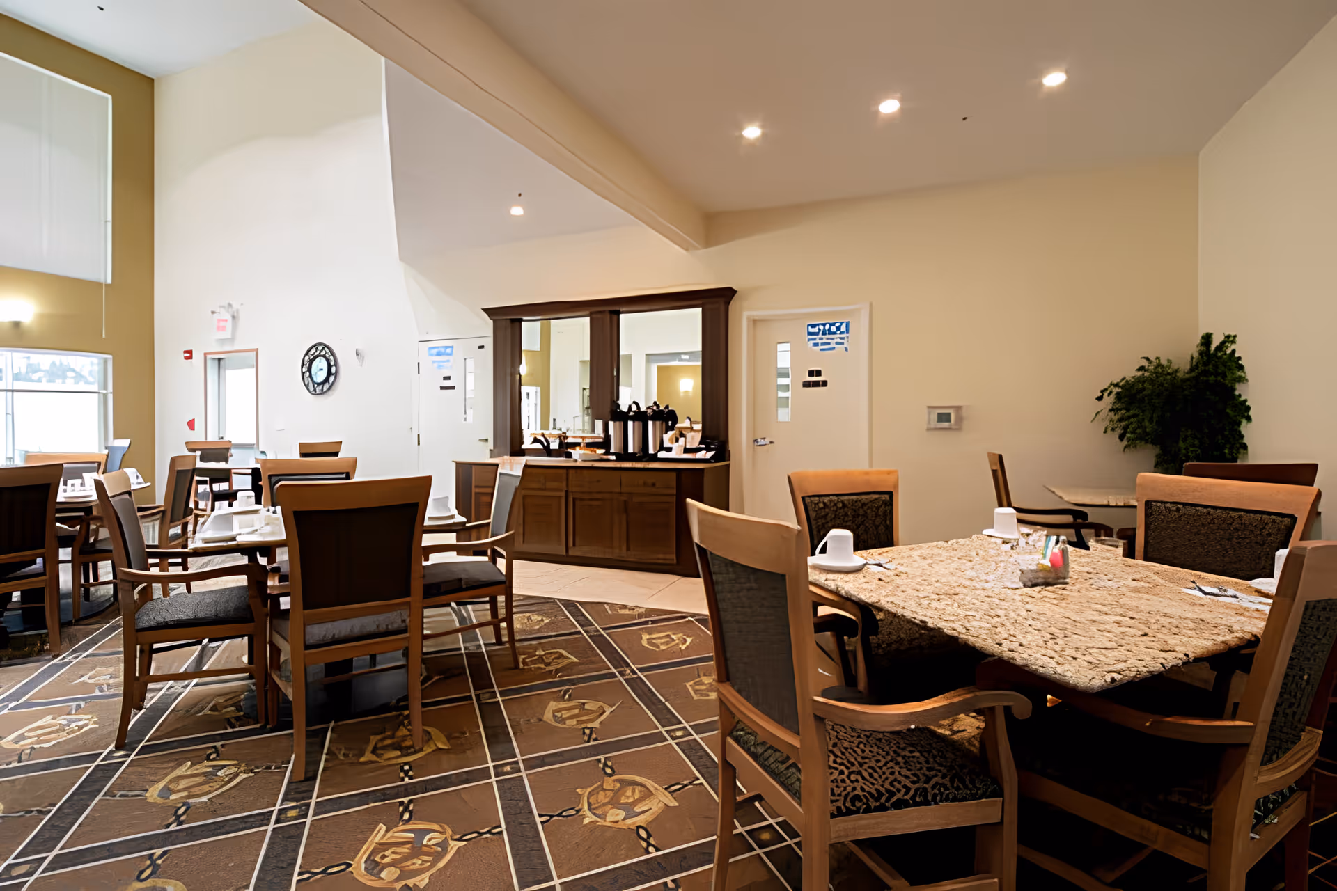A dining room in a senior living facility with several wooden tables and chairs arranged on a patterned carpet. The tables are set with white cups and utensils. There is a wooden buffet cabinet with coffee dispensers and a large mirror on the wall behind it. The room has beige walls and ceiling lights, with a large window letting in natural light.