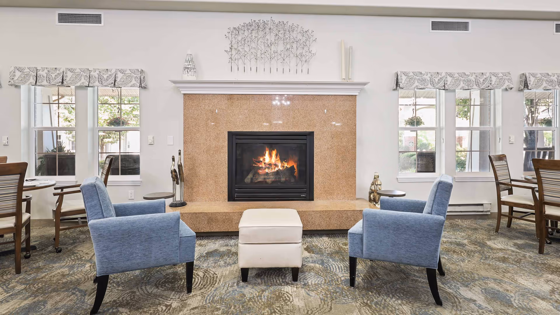 A cozy common area with a lit fireplace centered on a beige stone surround. Two blue upholstered armchairs face each other with a small white ottoman between them. On either side of the fireplace are small round tables with decorative sculptures. The room has large windows with patterned valances, letting in natural light. Wooden dining chairs and tables are visible on the left and right sides of the room.