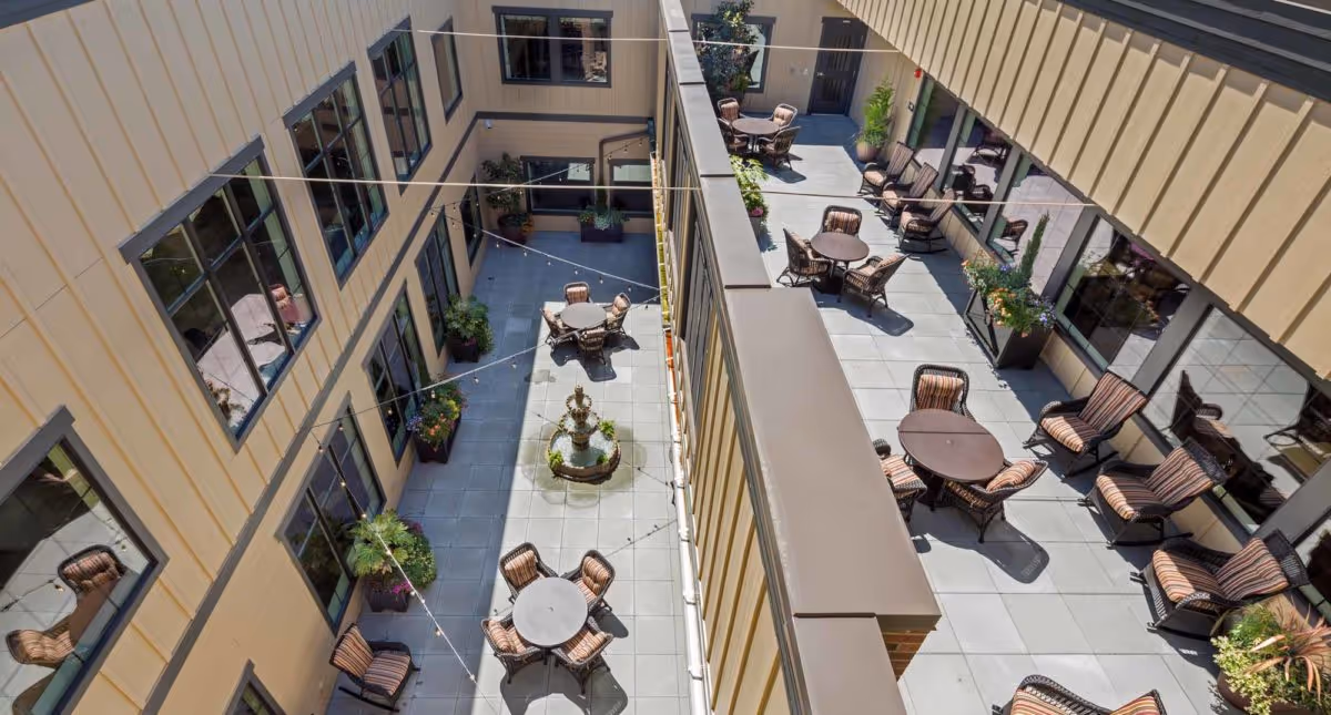 Aerial view of an outdoor courtyard area in a senior living facility with multiple round tables surrounded by cushioned chairs. The courtyard is enclosed by the building with large windows and decorated with potted plants and a central water fountain.