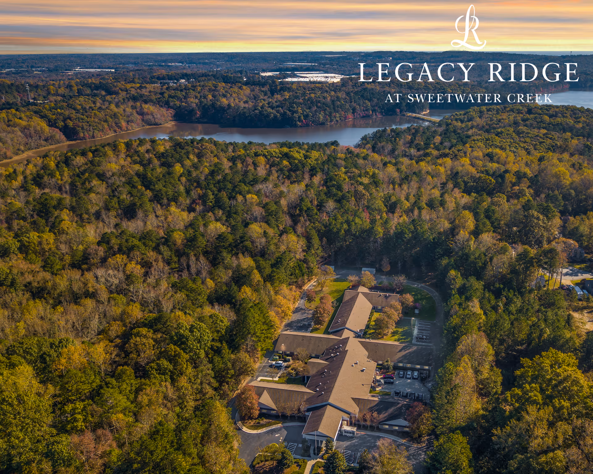 Aerial view of a senior living facility named Legacy Ridge at Sweetwater Creek surrounded by dense forest and a river in the background during sunset.