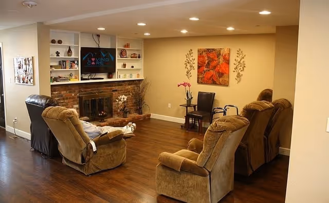 A cozy living room area with four brown recliner chairs arranged facing a brick fireplace and a wall-mounted TV. The TV screen displays the words 'Loving Care'. The room has wooden flooring, built-in white shelves with decorative items, and a small table with a pink orchid and a chair. The walls are beige with a large floral painting and decorative wall decals.