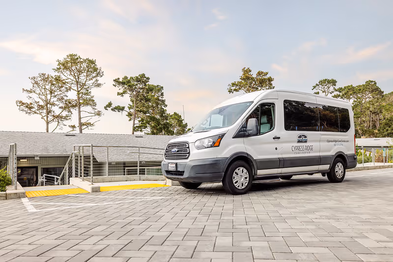 A white passenger van with Cypress Ridge Care Center branding is parked on a paved driveway in front of a building with a ramp and stairs. Trees and a partly cloudy sky are visible in the background.