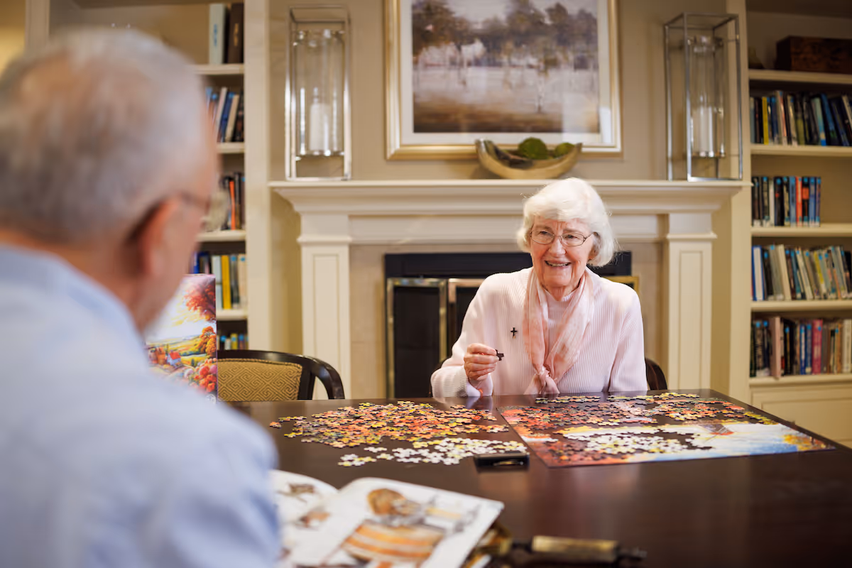 An elderly woman smiles while working on a jigsaw puzzle at a table in a cozy room with bookshelves and a fireplace while another person is partially visible.