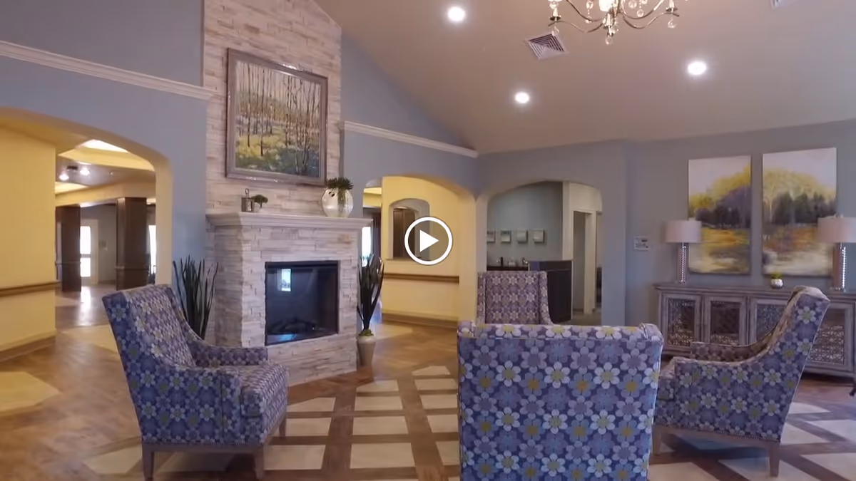 A cozy living room area with four patterned armchairs arranged around a modern stone fireplace. The room features wooden flooring with a geometric design, soft gray walls, a chandelier, and decorative paintings above a sideboard with two lamps.
