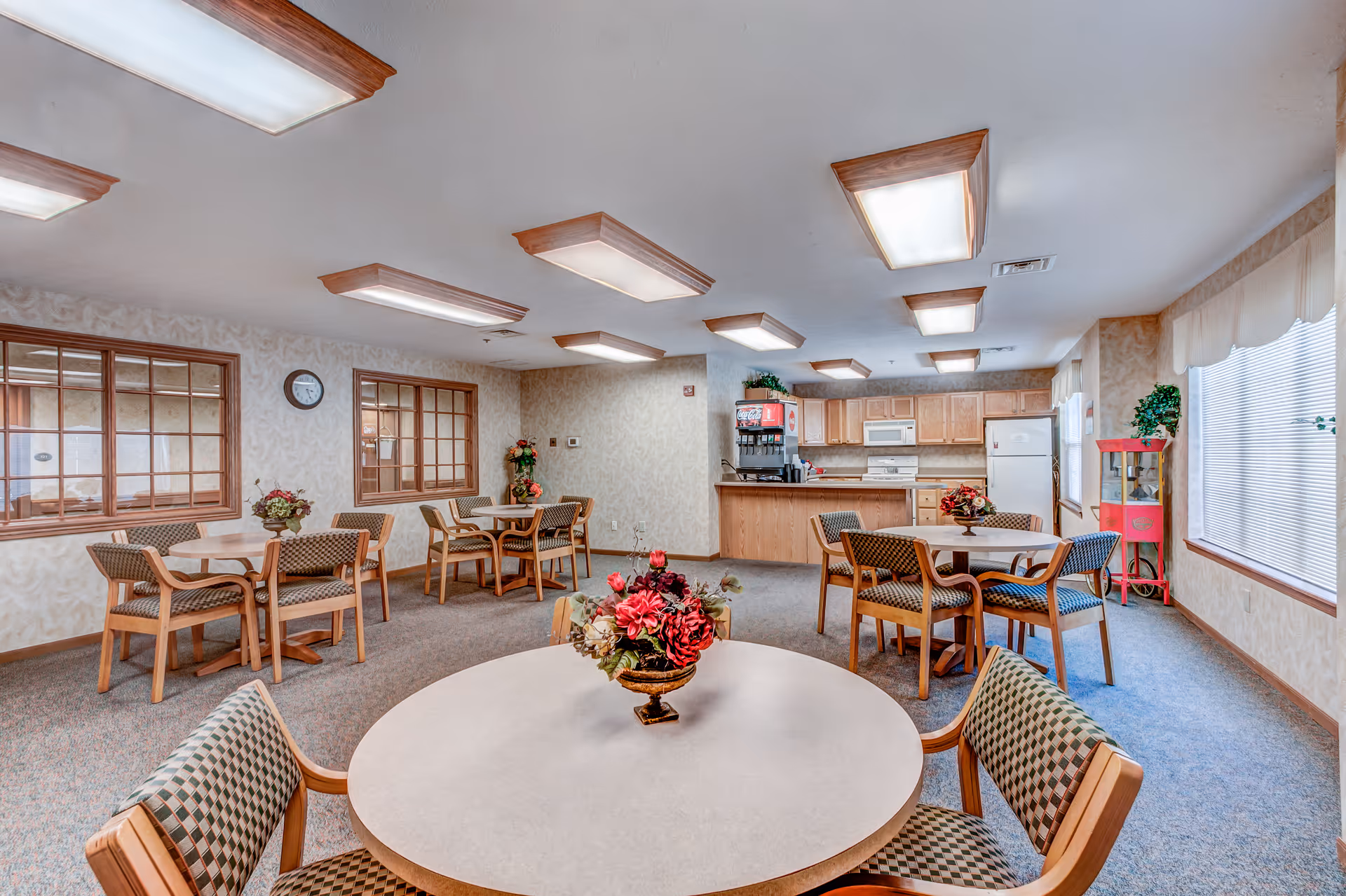 A bright dining room area in a senior living facility with multiple round tables and cushioned chairs arranged around them. Each table has a floral centerpiece. The room features large windows with blinds, a kitchen area with wooden cabinets, a refrigerator, microwave, stove, and a beverage dispenser. The ceiling has multiple rectangular light fixtures, and the walls are decorated with light-colored wallpaper and framed windows.