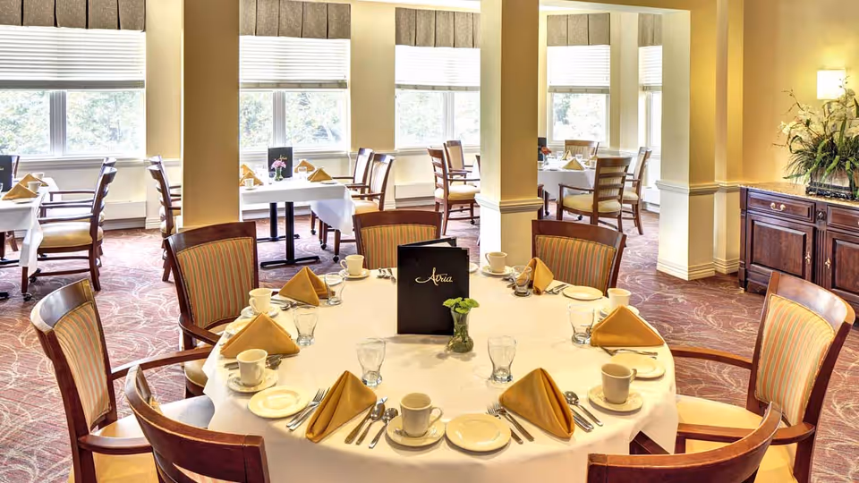 A bright dining room with round and rectangular tables set with white tablecloths, beige napkins, cups, plates, and silverware. The room has large windows with white blinds and beige walls. There is a dark wooden sideboard with a floral arrangement on the right side.