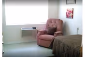 A small bedroom with a single bed covered in a brown quilt, a pink upholstered armchair with a dark pillow, a window with a white blind, a wall-mounted air conditioning unit, and a floral picture hanging on the wall.