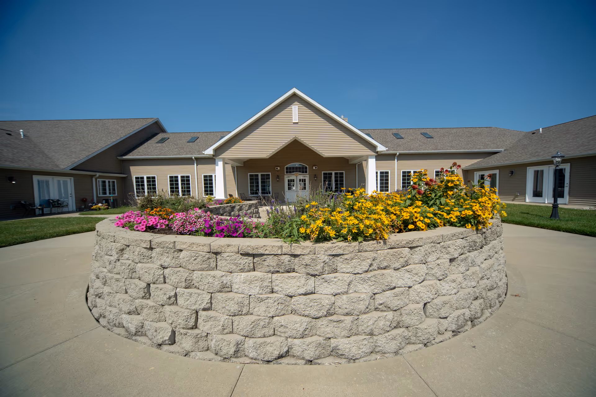Circular raised flower bed filled with colorful flowers in front of the entrance to a single-story senior living building under a clear blue sky.