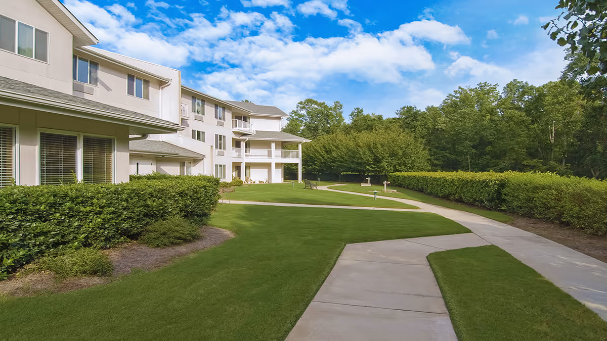 Exterior view of a multi-story senior living building with a curving concrete pathway, manicured lawn, and hedges under a blue sky.