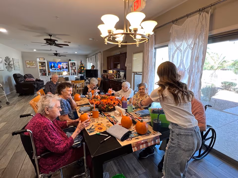 Older adults and a caregiver sit around a decorated dining table painting pumpkins in a bright assisted-living common room.