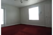 Empty room with white walls, a window letting in natural light, a closed white door, and dark red carpet flooring.