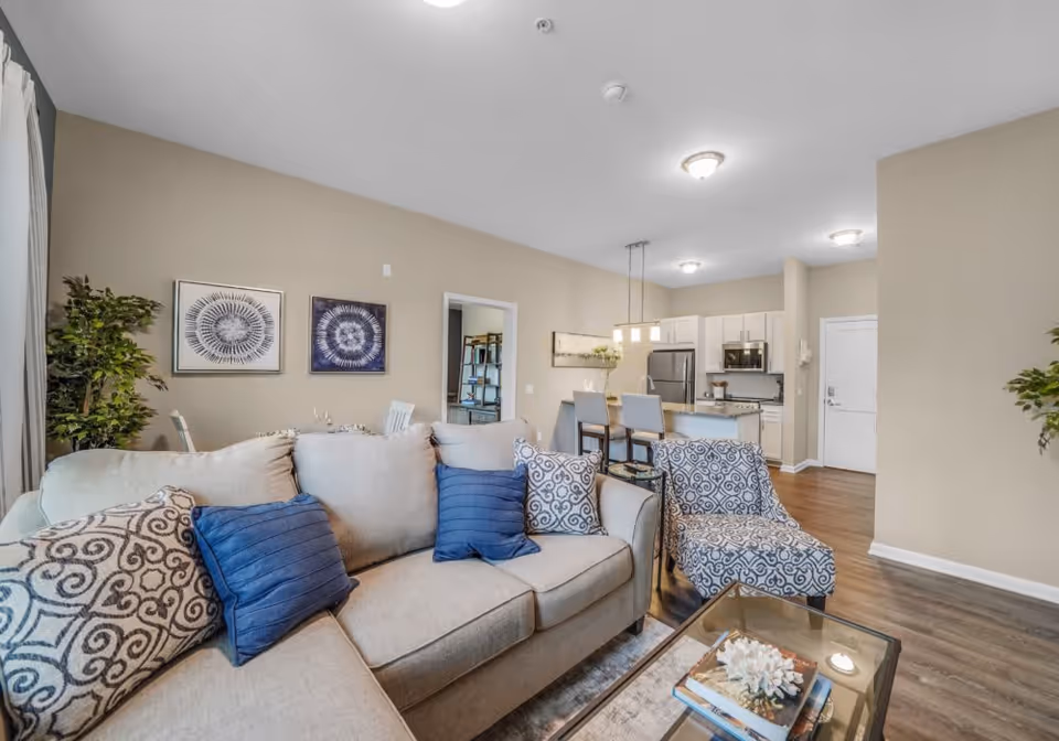 A modern living room and kitchen area in a senior living facility. The living room features a beige sofa with blue and patterned throw pillows, a patterned armchair, and a glass coffee table with books and decorative items. The kitchen in the background has white cabinets, stainless steel appliances, and a breakfast bar with three chairs. The walls are painted beige, and there are two framed artworks on the wall behind the sofa.