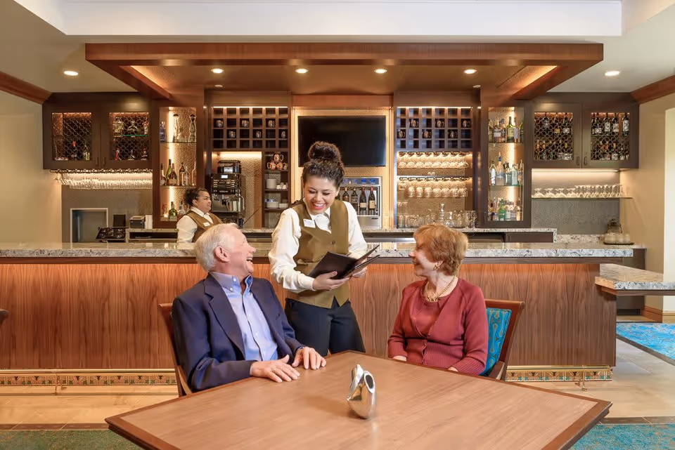 A server takes an order from two seated older adults at a table in a lounge-style dining area with a bar behind them.