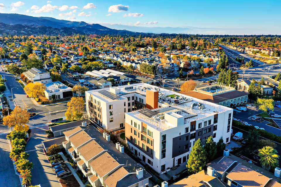 Aerial view of a multi-story assisted living building surrounded by residential streets, parking lots, and distant hills.