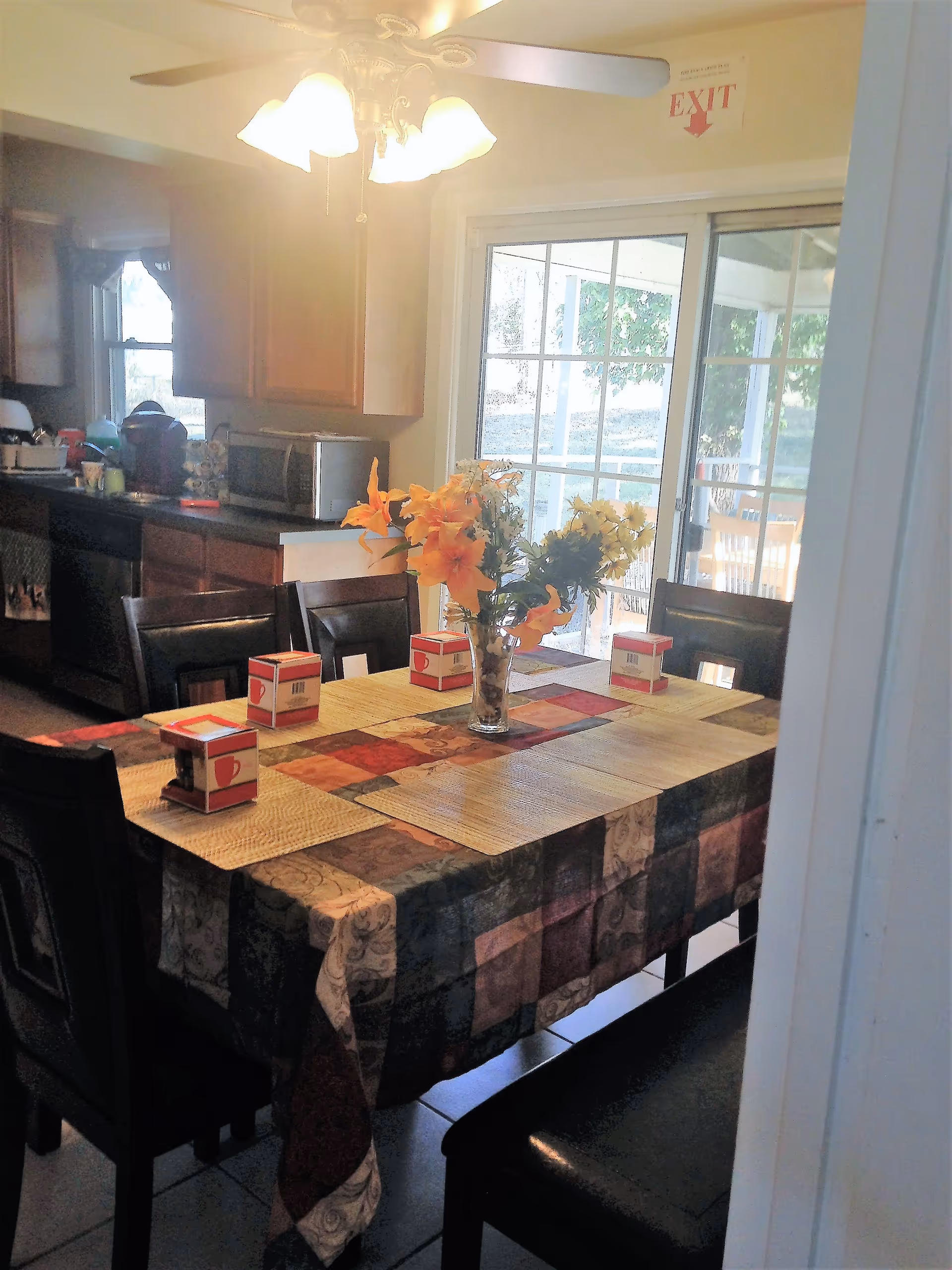 Dining room with a table set with placemats and a vase of flowers, surrounded by chairs and open to a kitchen with a sliding glass door.