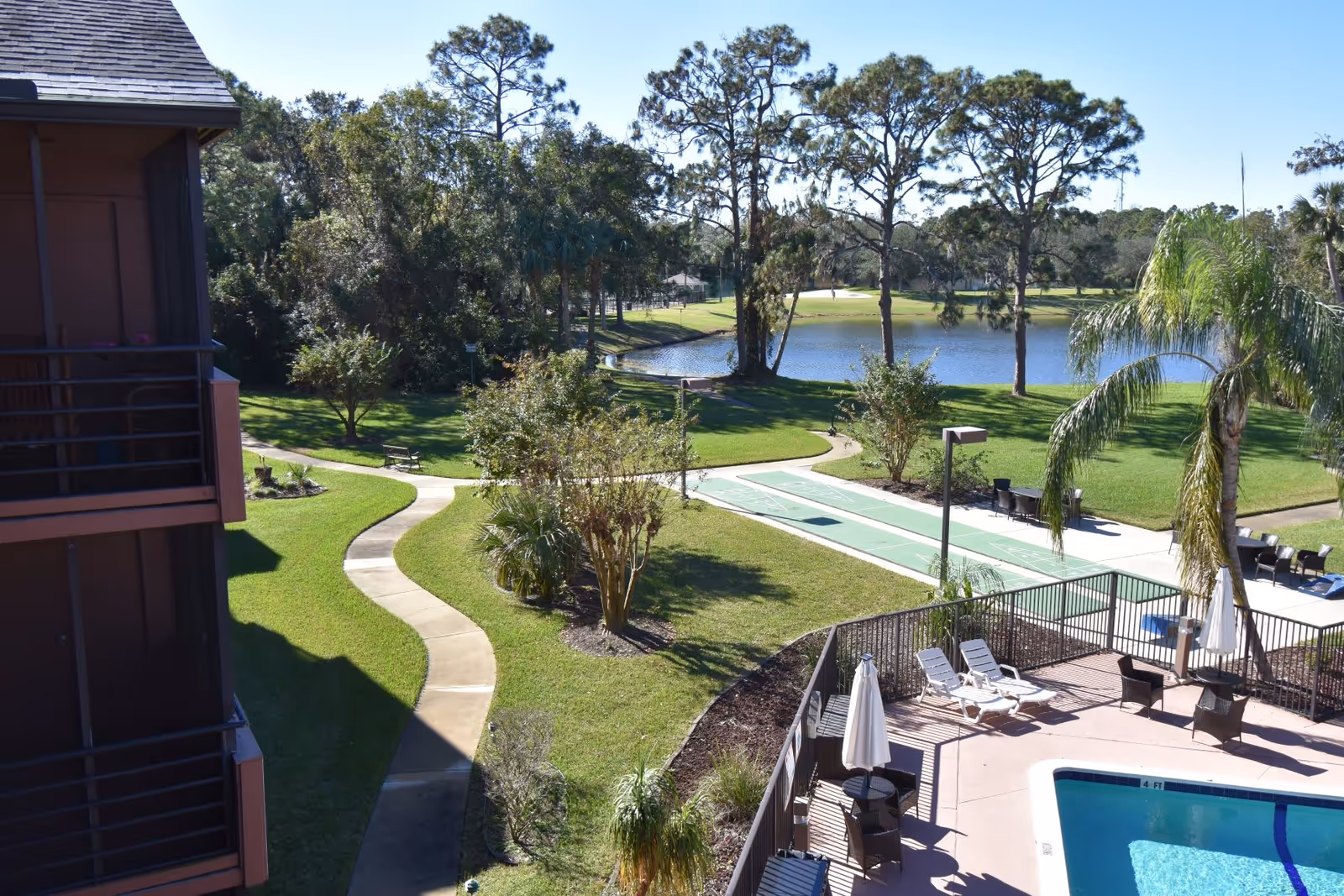 Outdoor view of a senior living facility showing a swimming pool area with lounge chairs and umbrellas, a shuffleboard court, paved walking paths, green lawns, trees, and a small lake in the background under a clear blue sky.